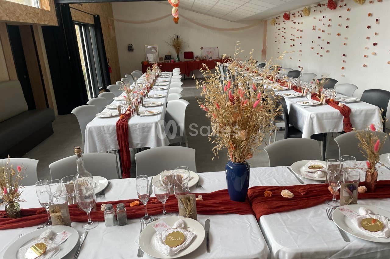 Salle décorée pour un événement avec tables alignées, nappes blanches, chemins de table rouges et décorations florales.
