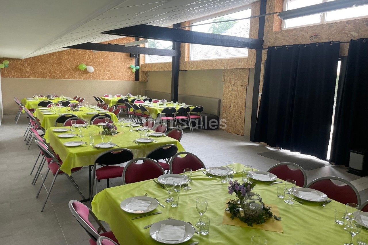 Salle décorée pour un événement avec des tables dressées, nappes vertes, chaises rouges et lumière naturelle.