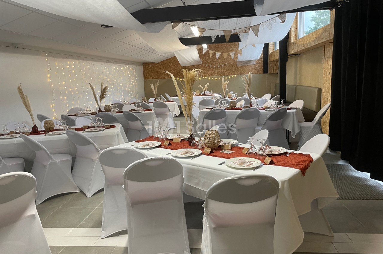Salle de réception décorée avec des tables dressées, nappes blanches, chemins de table rouges et éléments naturels.