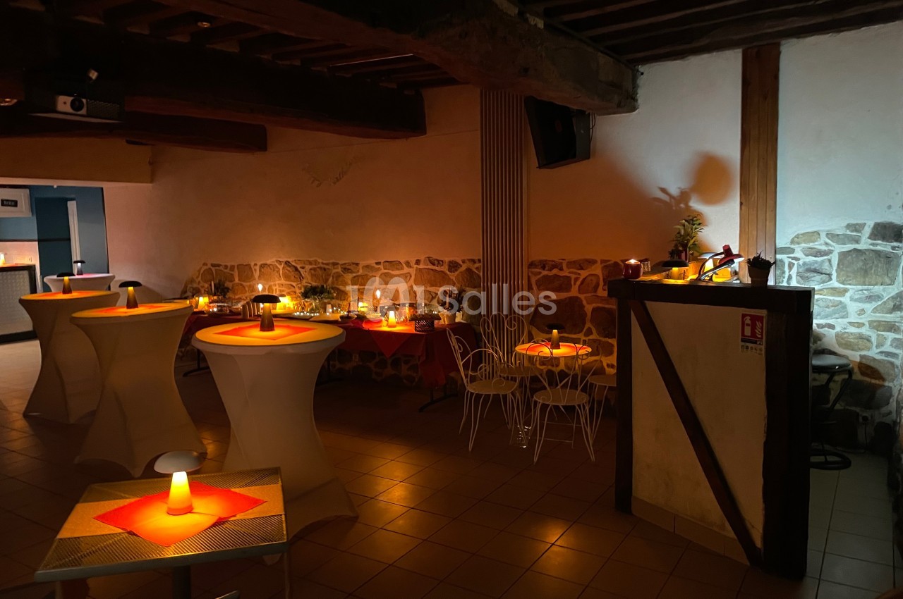 Salle faiblement éclairée avec des tables hautes et basses décorées de bougies, murs en pierre et poutres apparentes.
