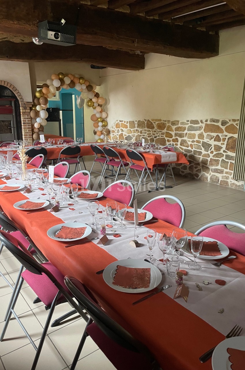 Salle décorée pour un repas festif avec longues tables dressées, nappes rouges, vaisselle et arche de ballons en arrière…