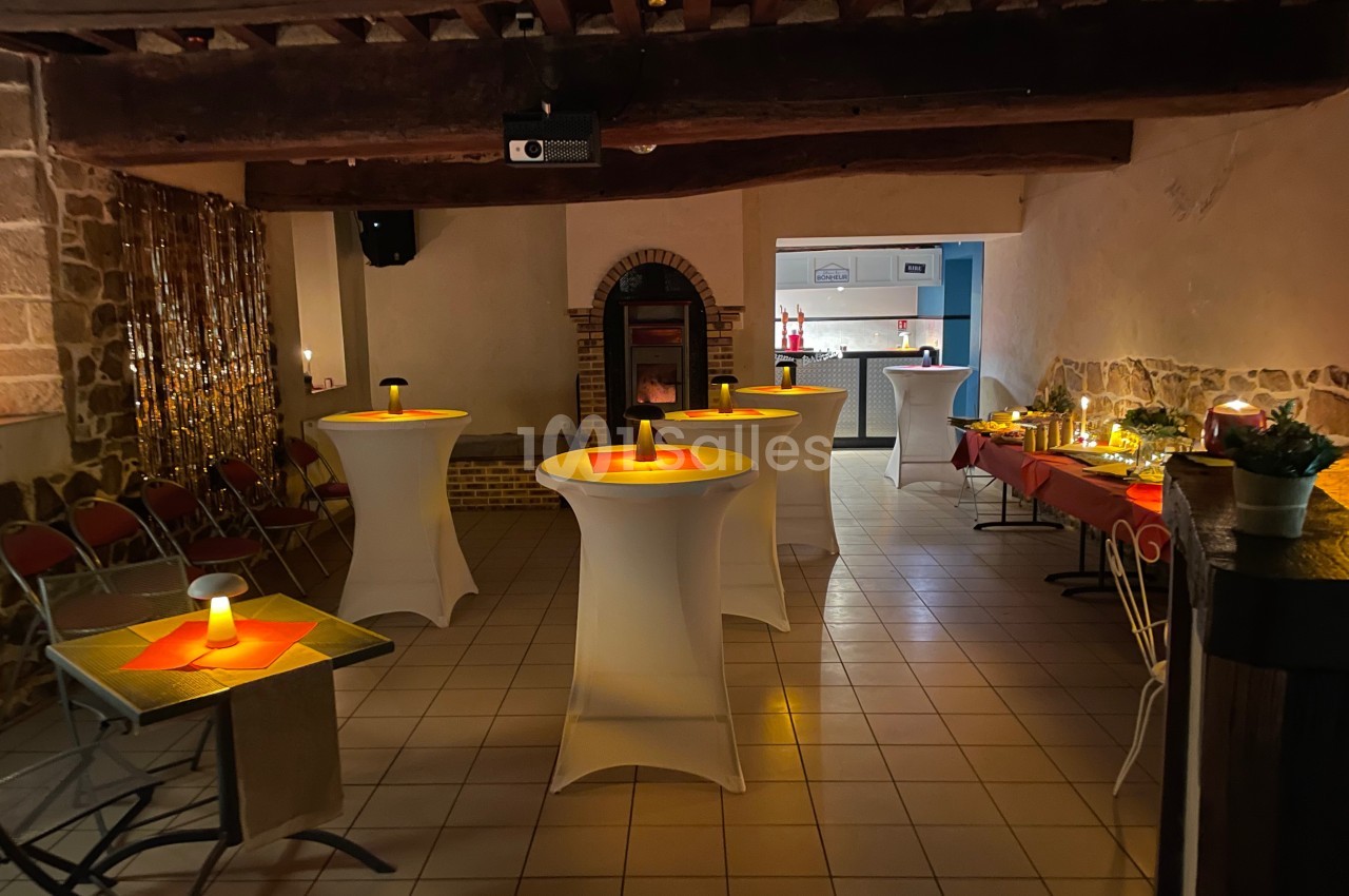 Salle décorée pour un événement, avec tables hautes, bougies et éclairage tamisé, dans un cadre rustique.
