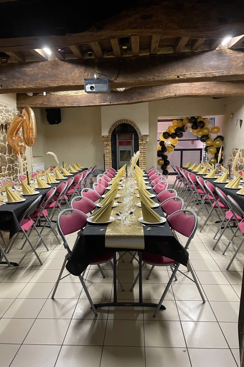 Salle décorée pour un événement avec tables alignées, nappes noires, chemins dorés, chaises roses et ballons en arrière-plan.