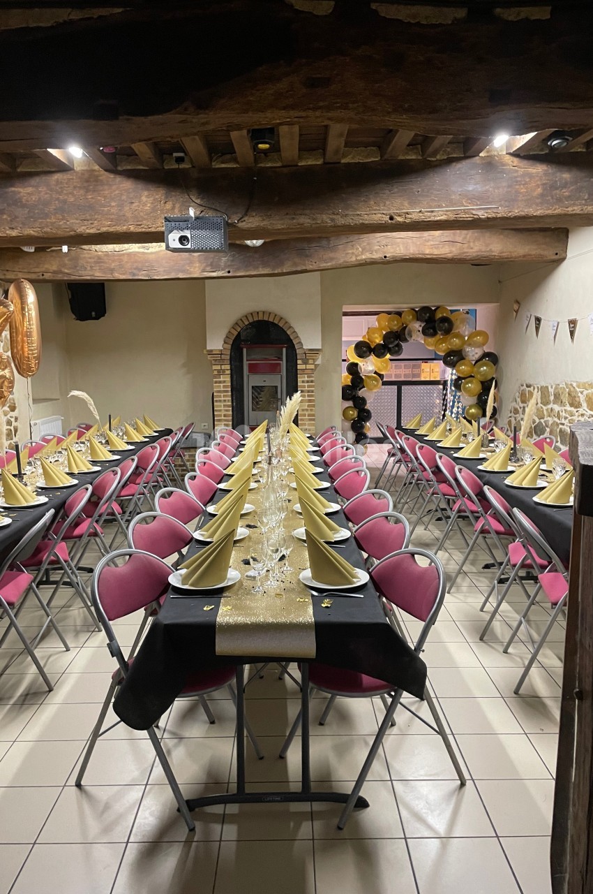Salle décorée pour un événement, avec tables alignées, nappes noires, serviettes dorées et ballons en arrière-plan.