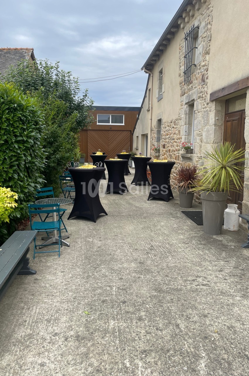 Cour extérieure avec tables hautes noires dressées, entourées de bancs, plantes et bâtiments en pierre.