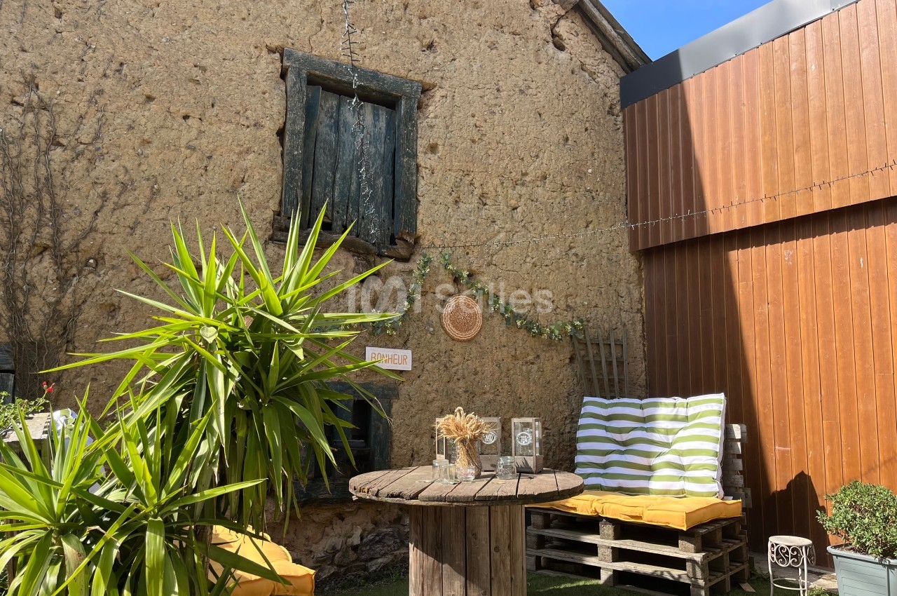 Cour extérieure avec mur en pierre, table en bois, coussins rayés sur palettes et plantes décoratives sous un ciel bleu.
