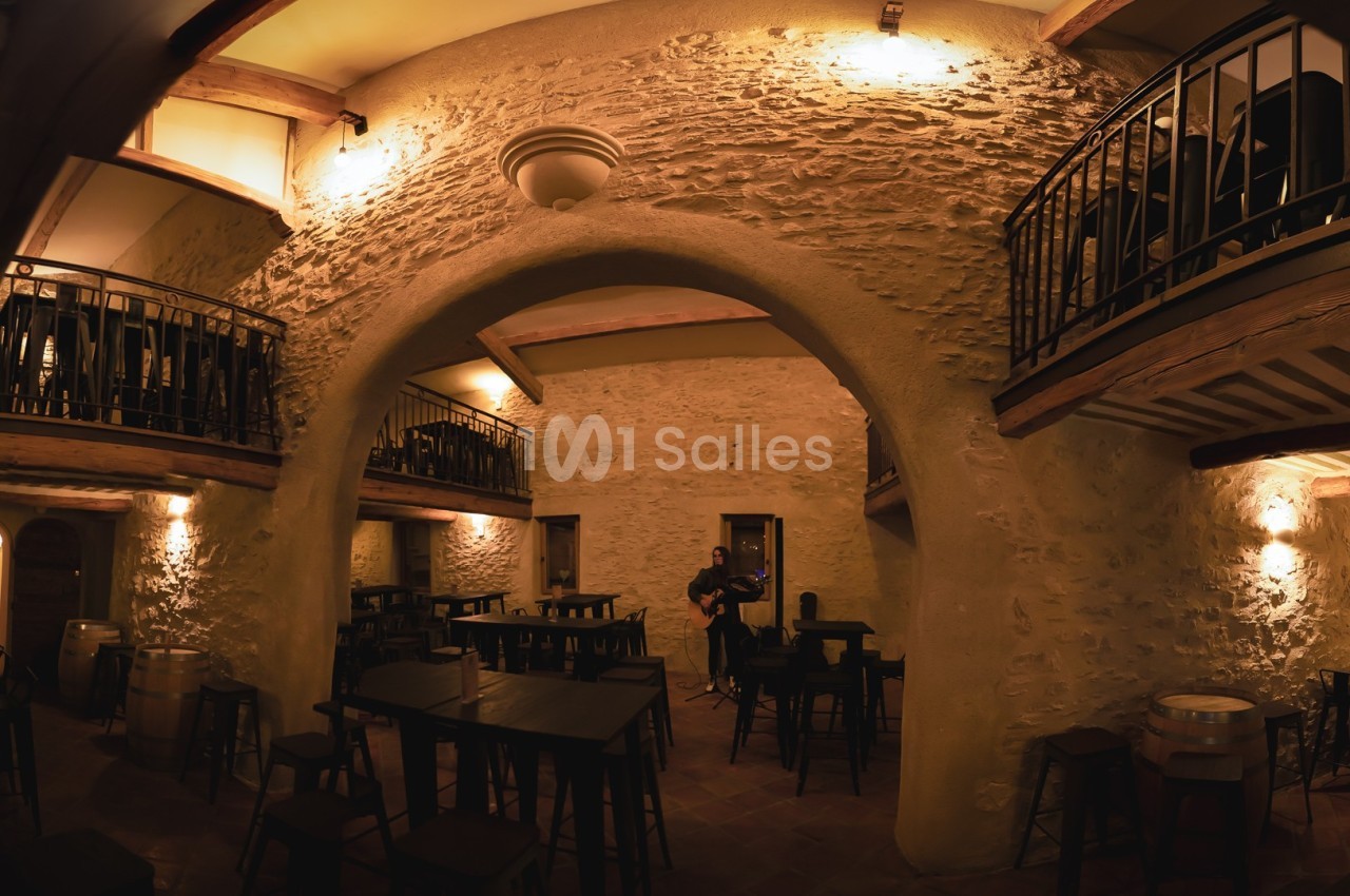 Salle intérieure en pierre avec arches, éclairage chaleureux, tables et chaises en bois, mezzanine avec balustrades.