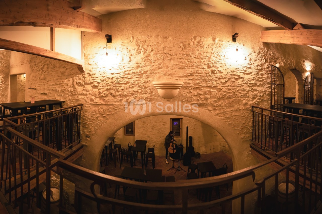 Salle intérieure en pierre avec mezzanine, éclairée par des lumières chaudes, tables et chaises disposées en bois sombre.