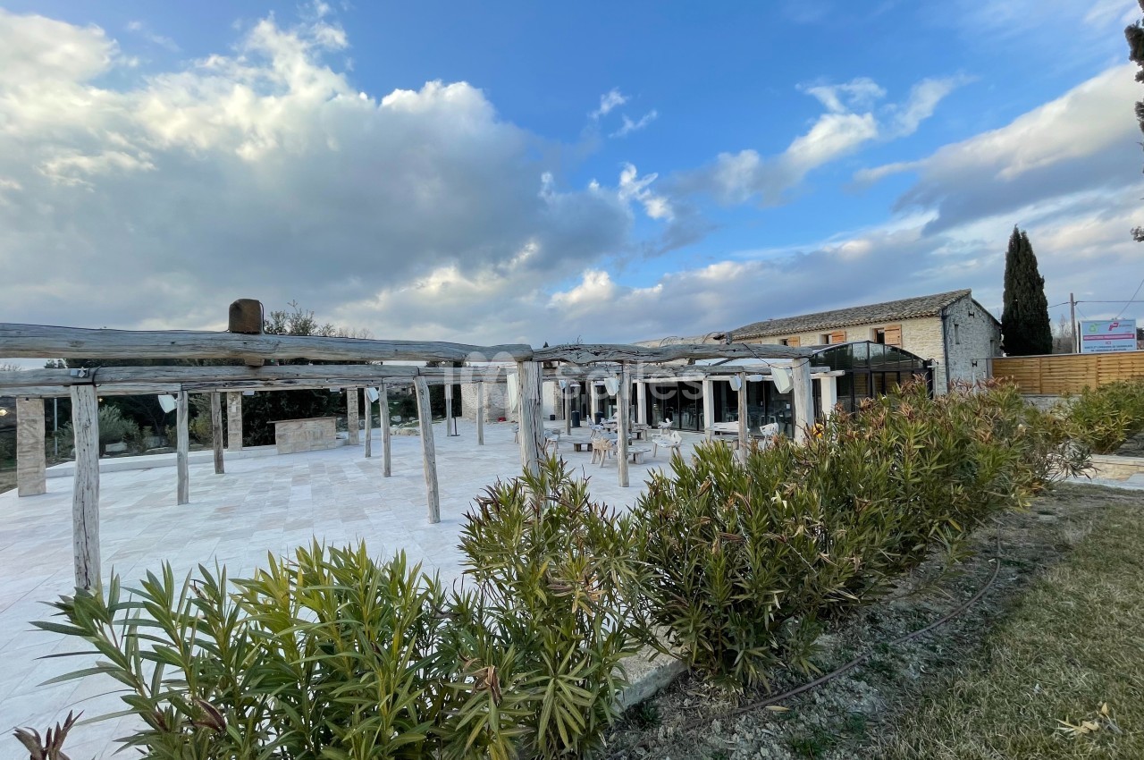 Cour extérieure avec pergolas en bois, entourée de végétation, devant un bâtiment en pierre sous un ciel partiellement…