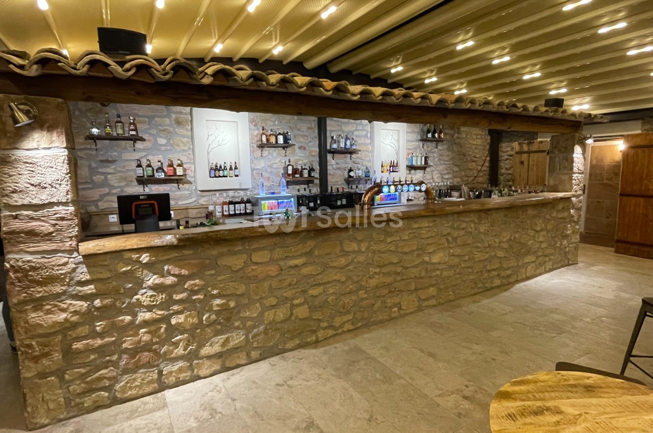 Comptoir de bar en pierre avec étagères garnies de bouteilles, éclairage chaleureux et plafond à poutres apparentes.