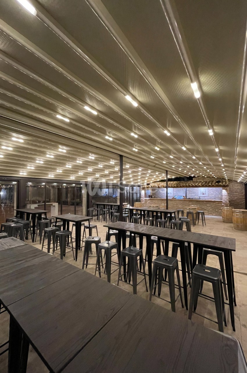 Salle spacieuse avec de longues tables en bois, chaises hautes et plafond éclairé par des rangées de lumières intégrées.