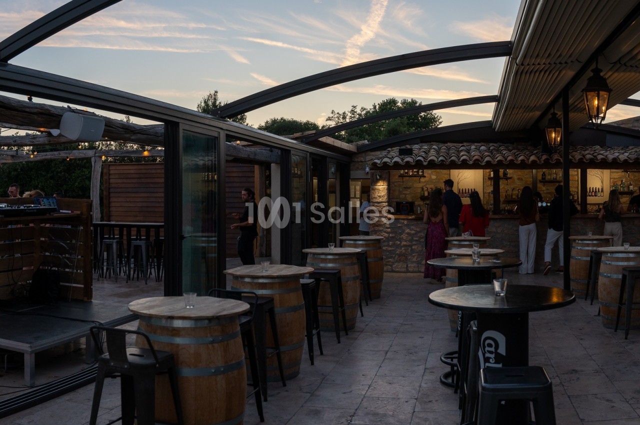 Terrasse extérieure avec tables en tonneaux, chaises hautes et toit partiellement ouvert, éclairée par une lumière douce.