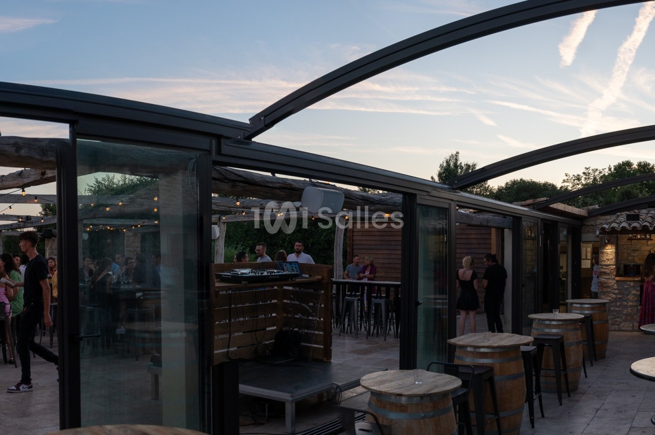 Terrasse couverte avec des tables et des chaises, des invités debout et assis, au coucher du soleil.