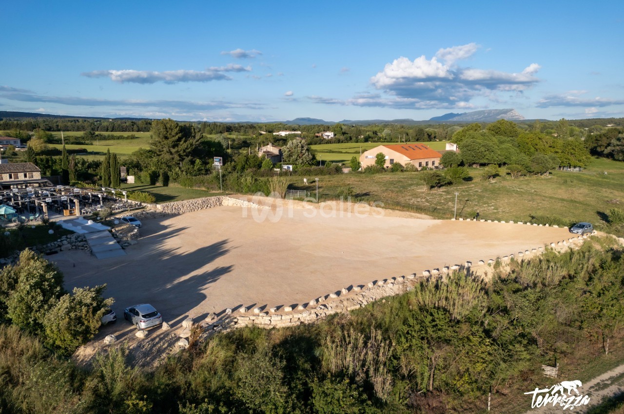Vue aérienne d'un grand parking en gravier entouré de verdure, avec des bâtiments et des collines à l'arrière-plan.