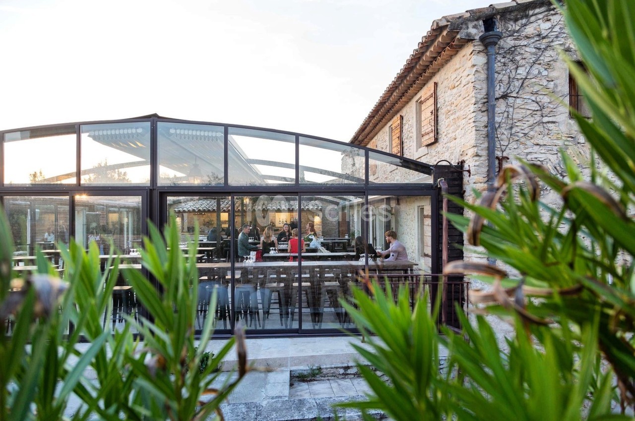 Salle de restaurant lumineuse avec verrière, tables dressées et clients attablés, entourée de végétation.