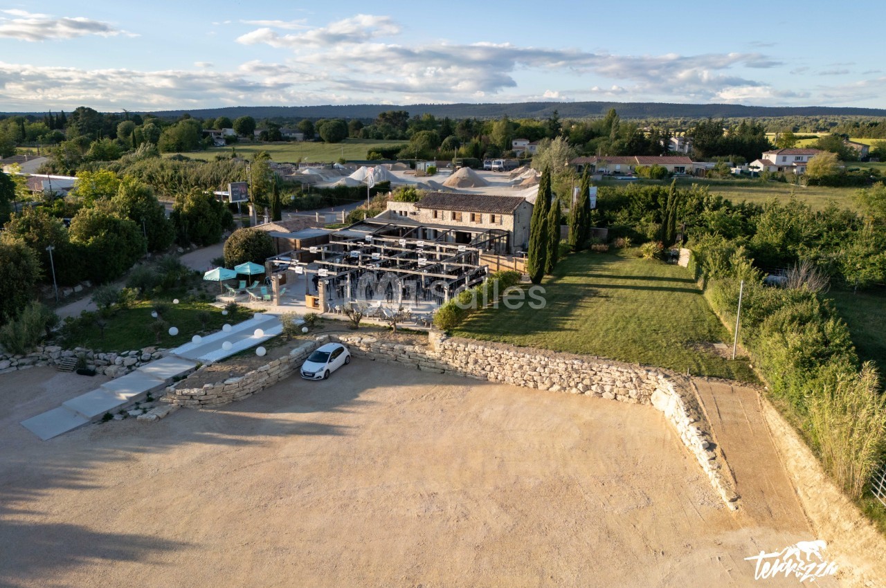 Vue aérienne d'un bâtiment en pierre avec une terrasse aménagée, entouré de verdure et d'un parking en gravier.