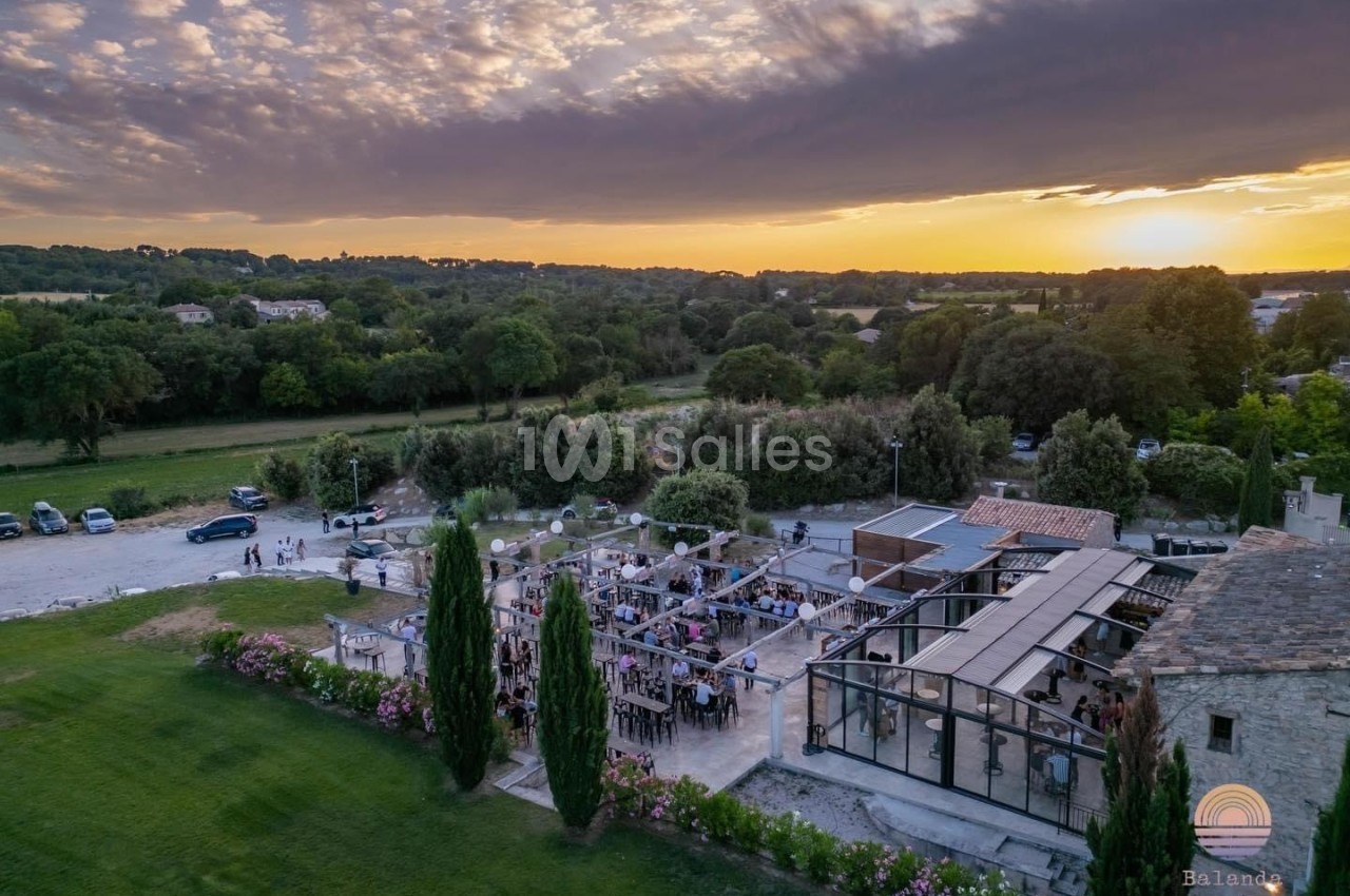 Vue aérienne d'une terrasse animée entourée de verdure au coucher du soleil, avec un ciel partiellement nuageux.