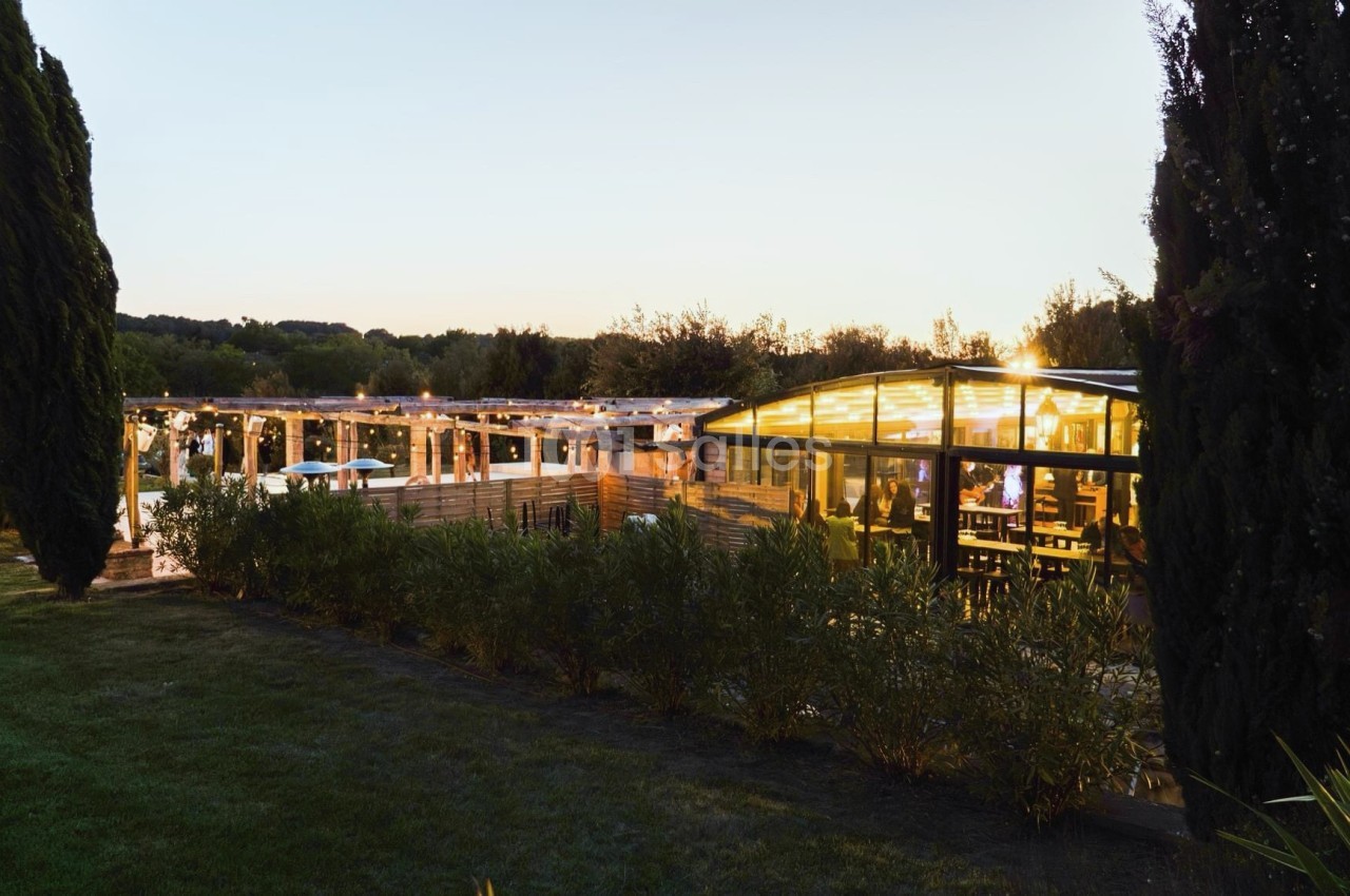 Vue d'un restaurant éclairé en soirée, entouré de végétation et d'arbres, avec une terrasse illuminée.