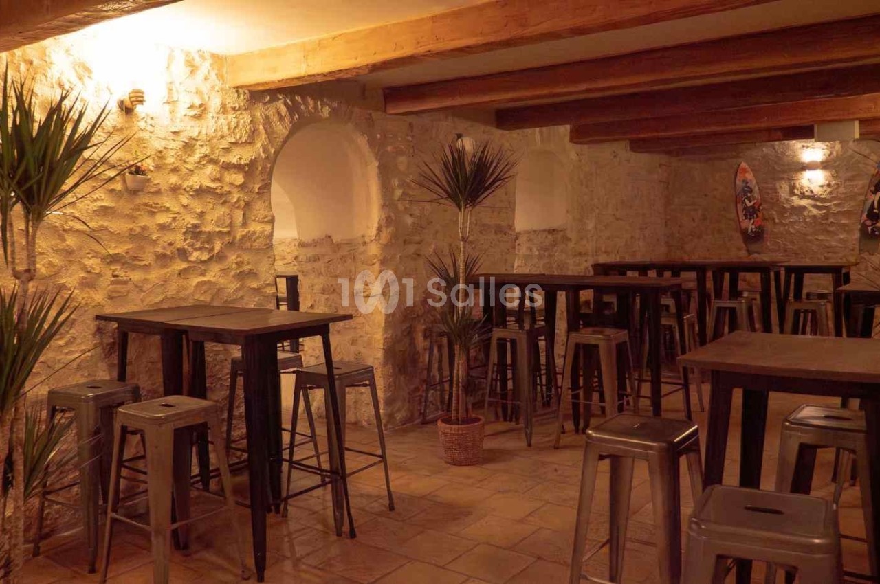 Salle avec murs en pierre, tables hautes en bois, tabourets métalliques et plantes décoratives sous un éclairage chaleureux.