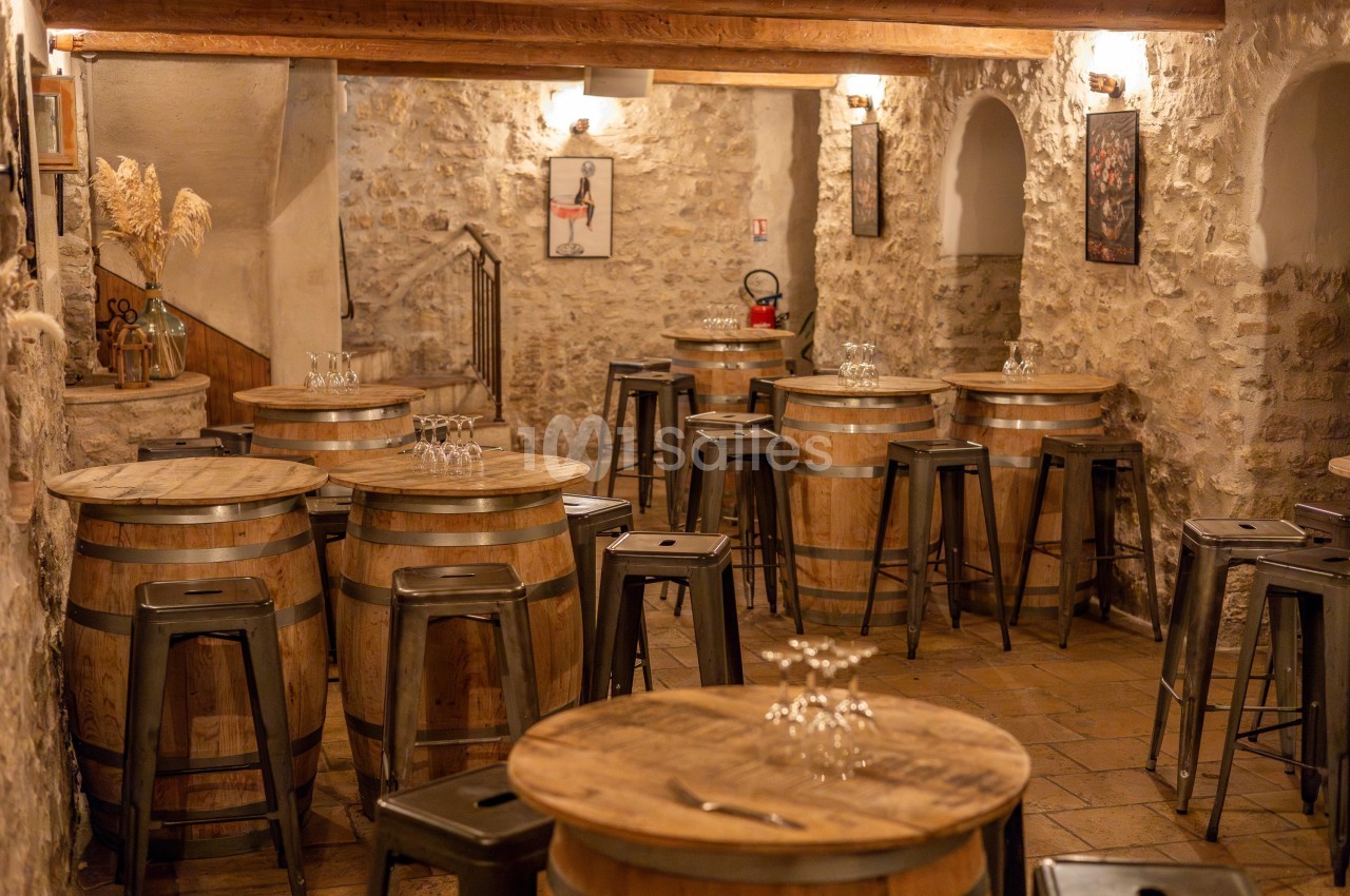 Salle rustique avec murs en pierre, tonneaux en bois servant de tables, tabourets métalliques et éclairage chaleureux.