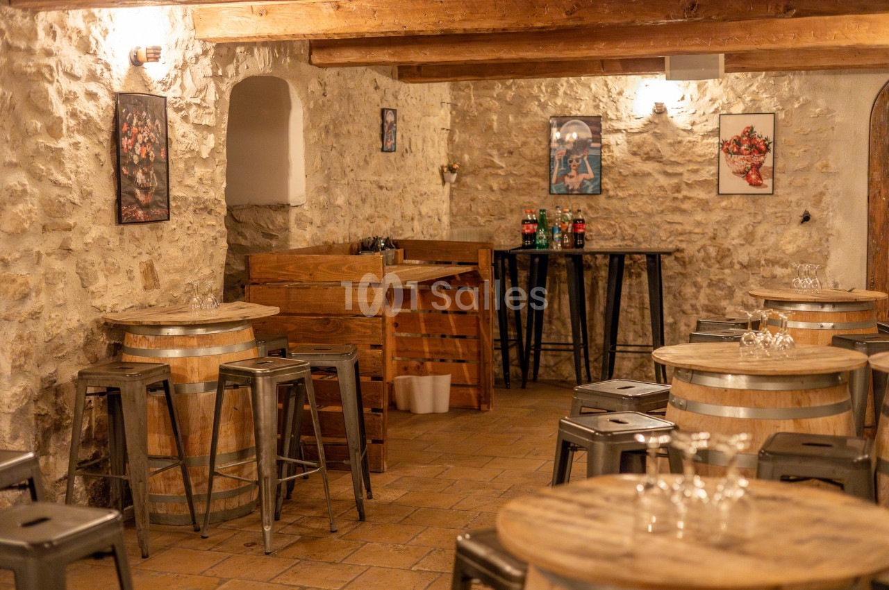 Salle rustique avec murs en pierre, tonneaux en bois servant de tables, tabourets métalliques et éclairage chaleureux.