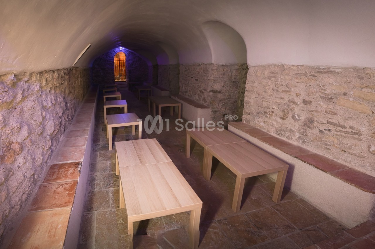 Salle voûtée en pierre avec des bancs et des tables en bois, éclairée par une lumière colorée provenant d'une fenêtre.