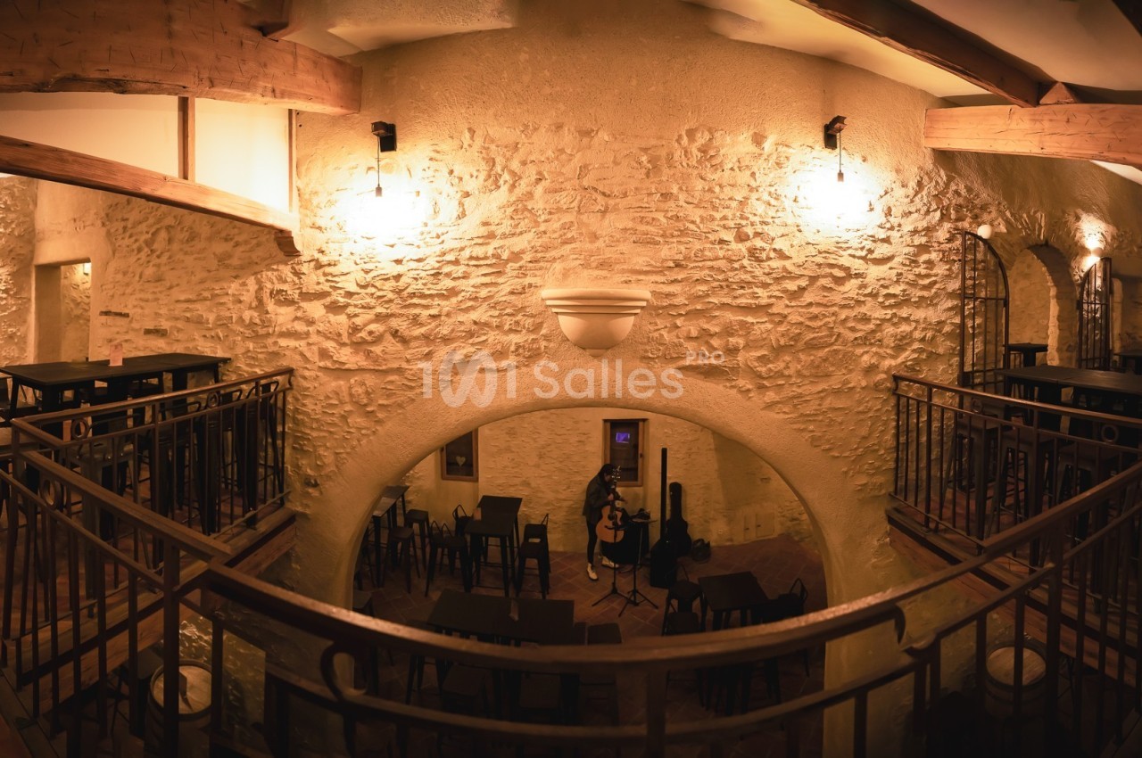 Salle intérieure en pierre avec mezzanine, éclairage chaleureux, tables hautes et chaises disposées autour.