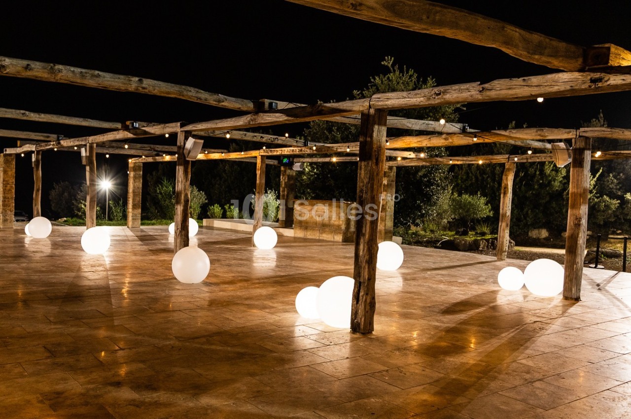 Structure en bois éclairée la nuit, avec des sphères lumineuses blanches posées sur un sol en pierre.