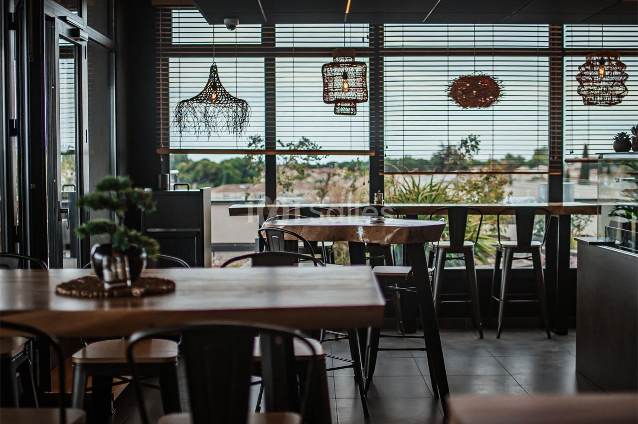 Salle de restaurant moderne avec tables en bois, chaises noires et grandes fenêtres donnant sur un paysage extérieur.