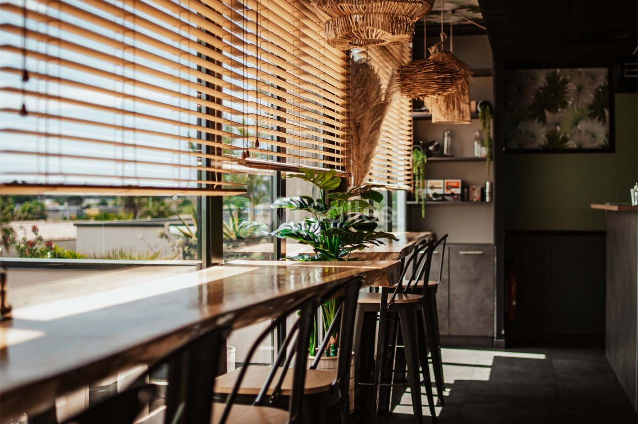 Salle lumineuse avec des tables en bois, des chaises hautes, des plantes et des stores laissant passer la lumière naturelle.