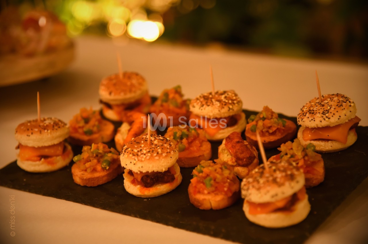 Plateau de mini-burgers et amuse-bouches variés disposés sur une ardoise, éclairage chaleureux en arrière-plan.