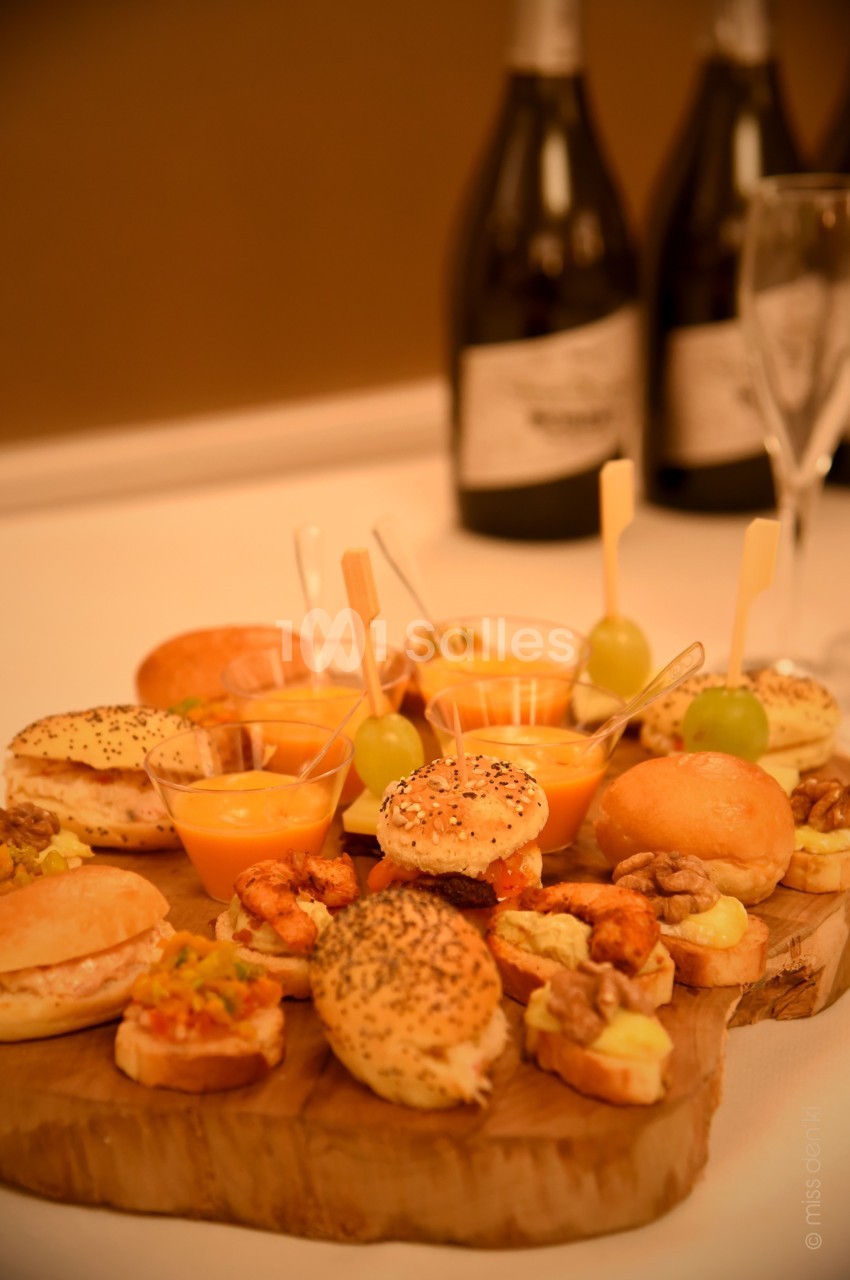 Plateau en bois garni de mini-burgers, verrines, toasts et amuse-bouches, avec deux bouteilles en arrière-plan.