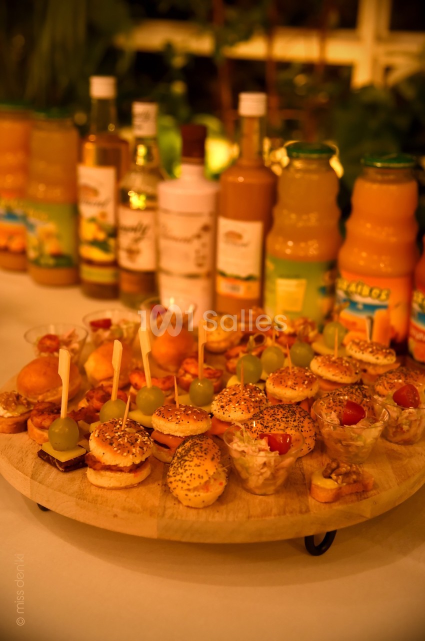 Plateau en bois garni de mini-burgers, verrines et amuse-bouches, avec des bouteilles de jus en arrière-plan.