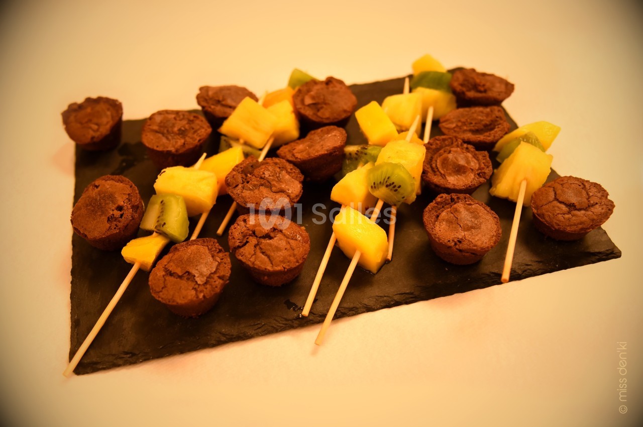 Brochettes composées de mini moelleux au chocolat, morceaux de kiwi et d'ananas, présentées sur une ardoise noire.