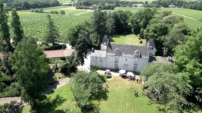 Vue aérienne d'un manoir entouré de jardins verdoyants et de vignobles, avec des personnes rassemblées sur la pelouse.