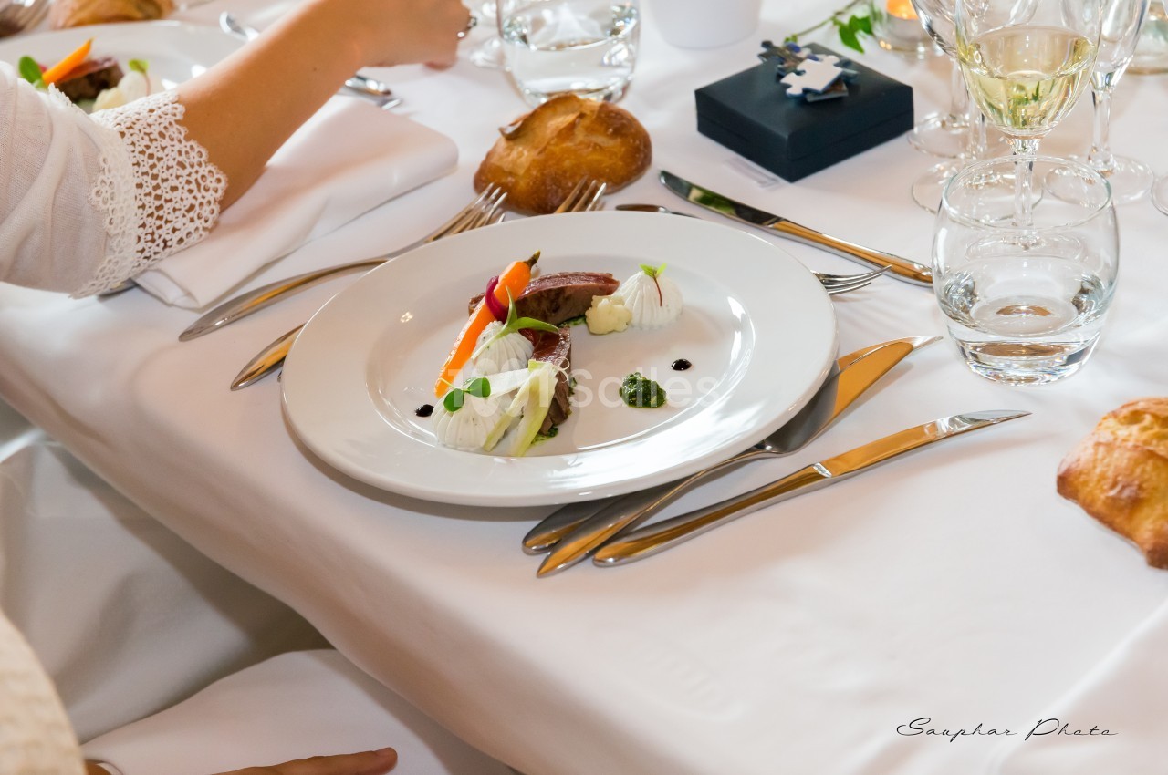 Assiette élégamment dressée avec légumes, viande et sauce, sur une table blanche avec couverts et verres.