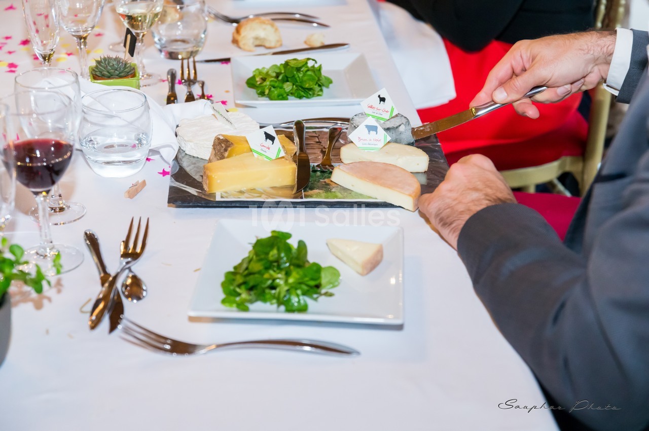 Une personne sert du fromage varié sur une assiette lors d'un repas, avec des salades et des verres sur la table.