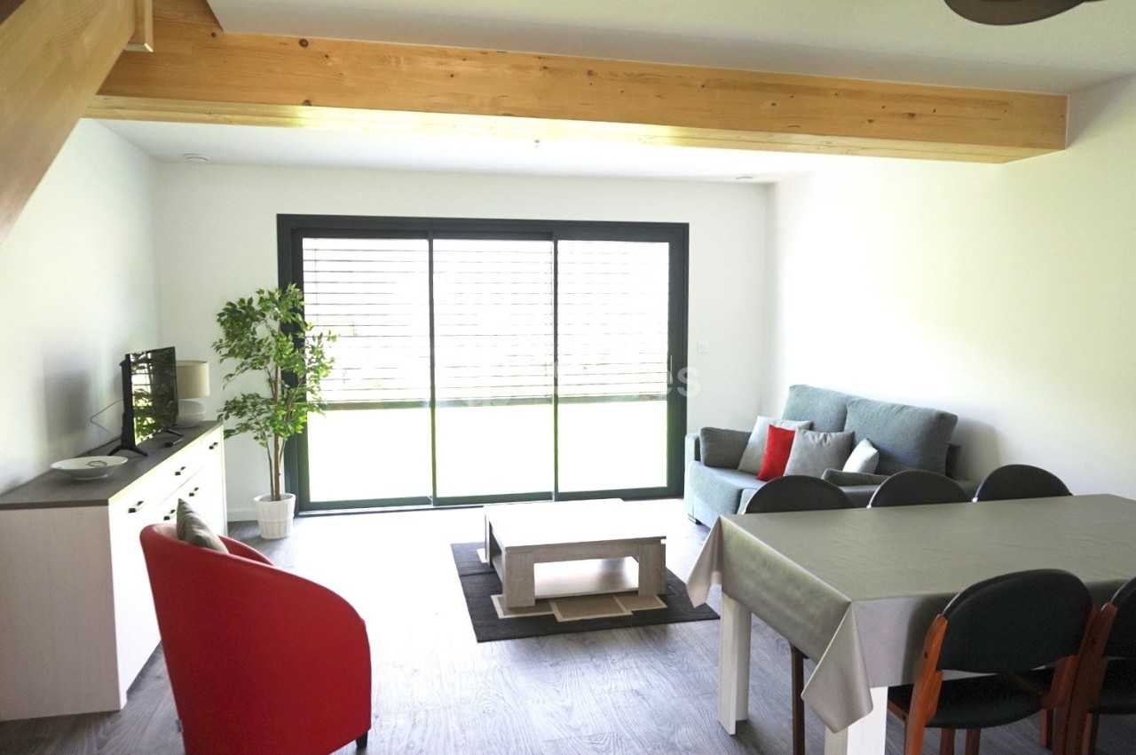 Salon lumineux avec canapé, table à manger, fauteuil rouge, meuble TV, plante verte et baie vitrée donnant sur l'extérieur.