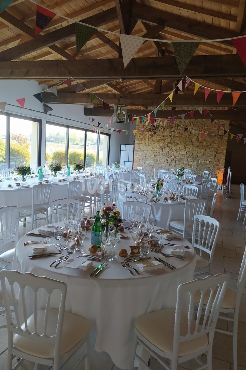 Salle de réception décorée avec des tables rondes dressées, des guirlandes colorées et une grande baie vitrée.