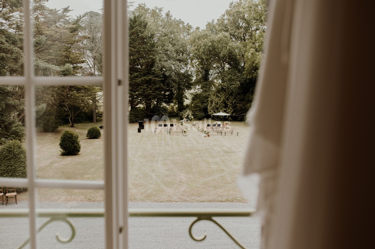 Vue depuis une fenêtre ouverte sur un jardin avec des chaises disposées pour un événement en plein air.