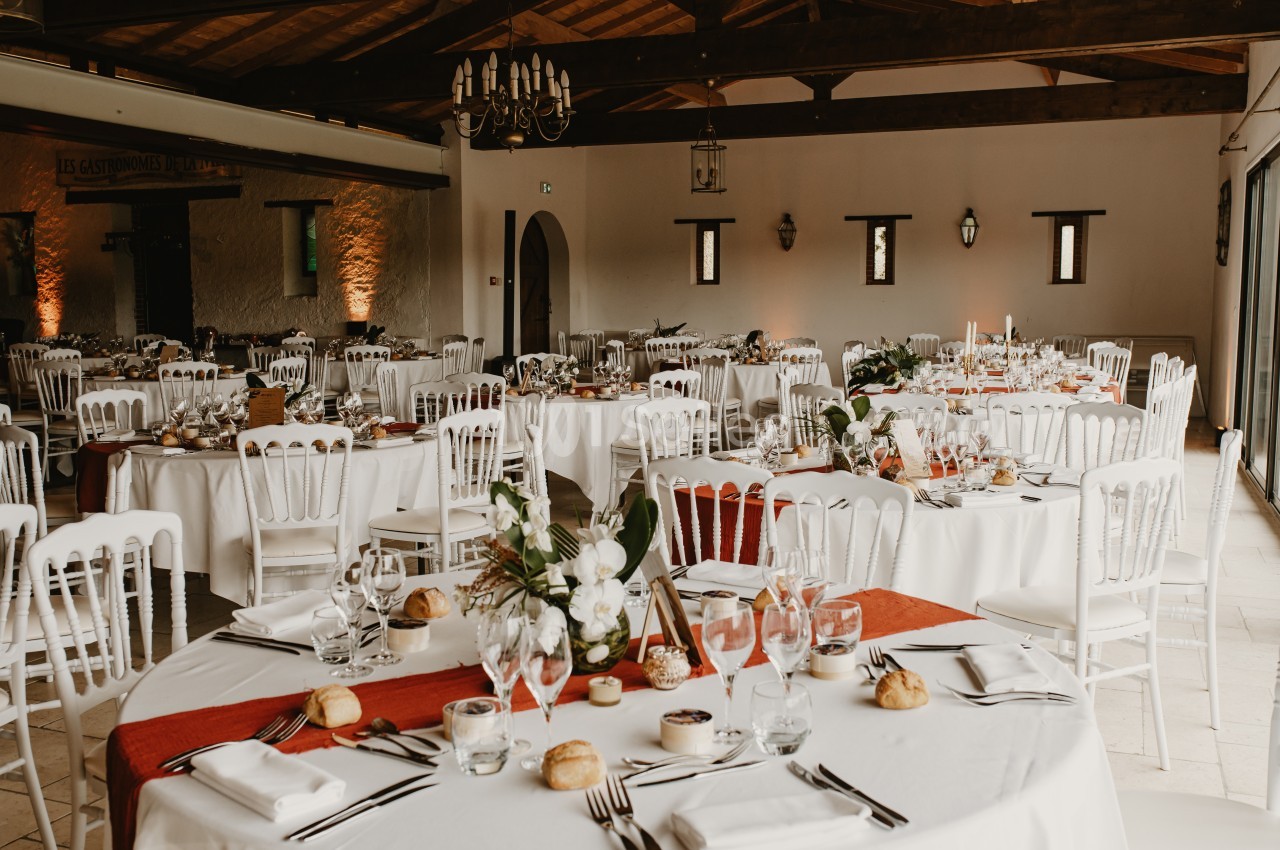 Salle de réception décorée avec des tables rondes dressées, nappes blanches, chaises blanches et arrangements floraux.