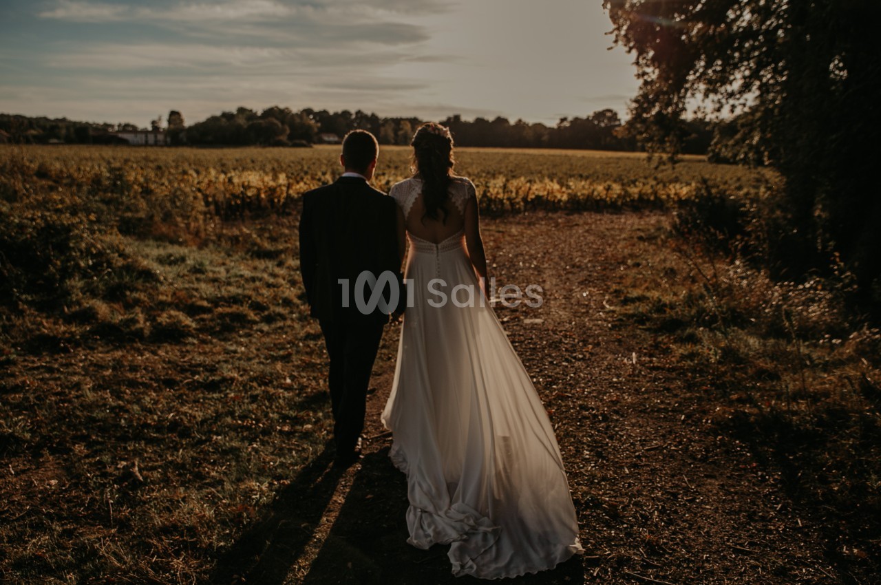 Un couple en tenue de mariage marche sur un chemin rural au coucher du soleil, entouré de végétation.