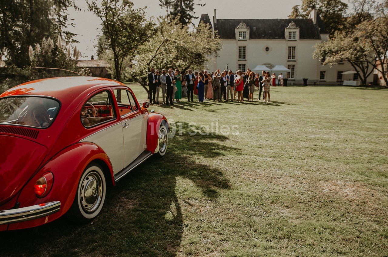 Une voiture rouge est garée sur une pelouse devant un groupe de personnes rassemblées près d'un bâtiment ancien.