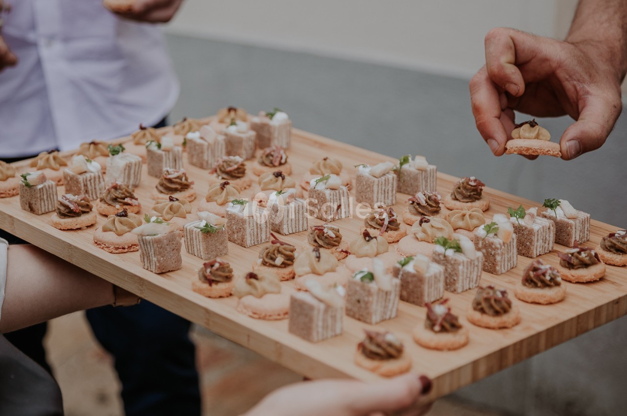 Plateau en bois garni de canapés variés, tenu par plusieurs mains, avec une personne prenant un amuse-bouche.