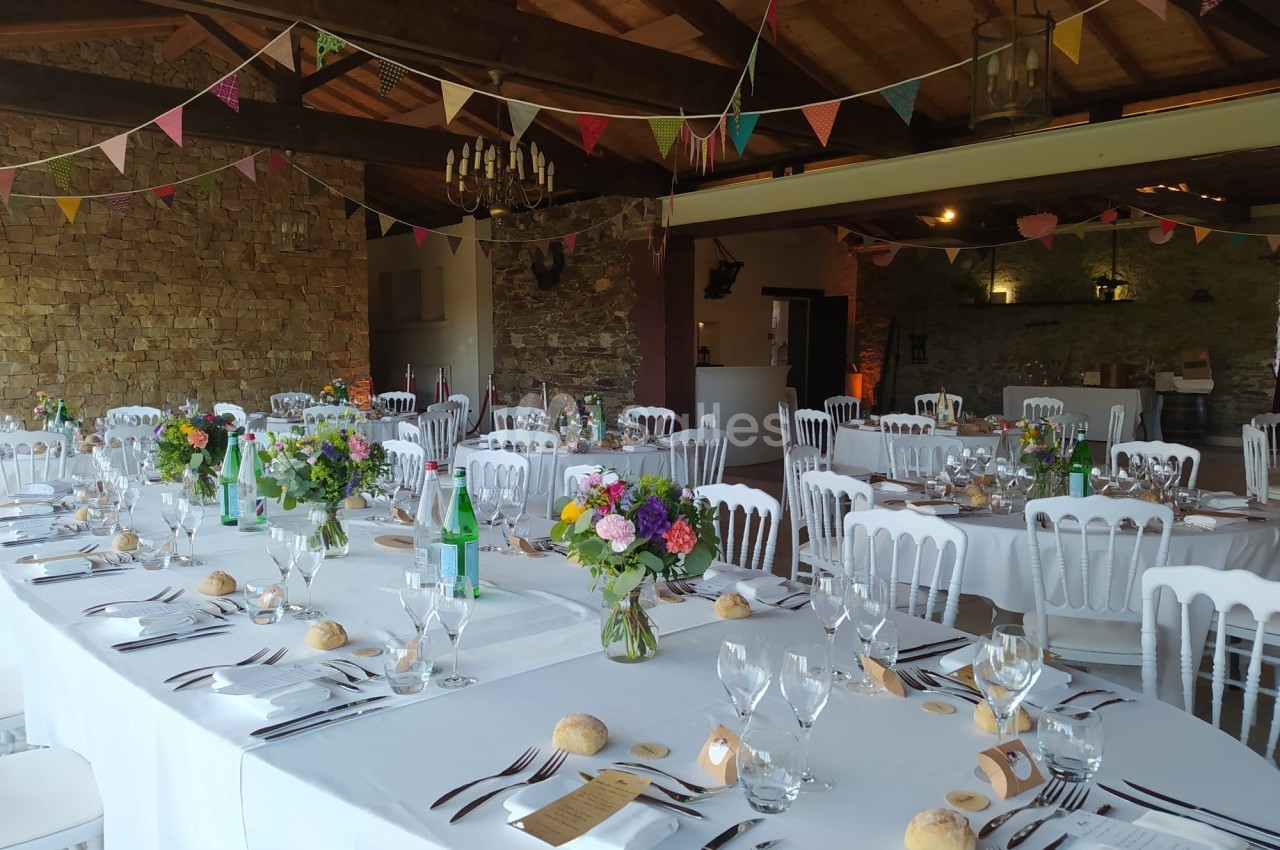 Salle de réception décorée avec des tables dressées, nappes blanches, fleurs colorées et guirlandes suspendues.