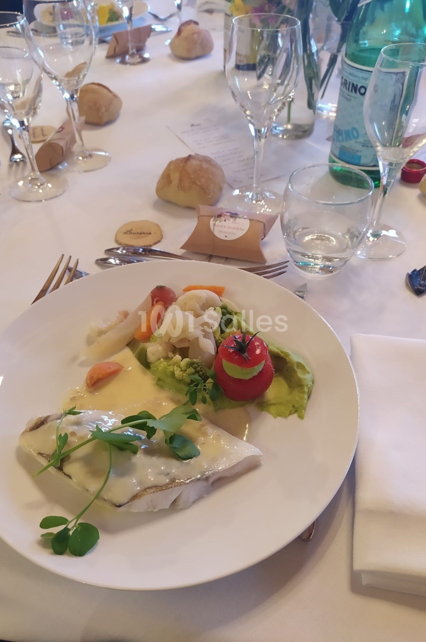 Assiette de poisson avec sauce, légumes variés, purée verte, et décoration, sur une table dressée pour un repas.