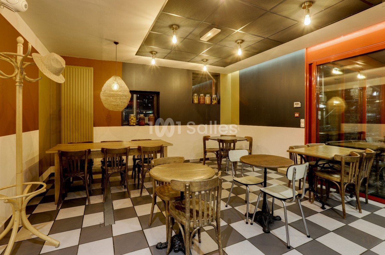 Salle de restaurant avec tables en bois, chaises dépareillées, éclairage suspendu et sol à carreaux noir et blanc.