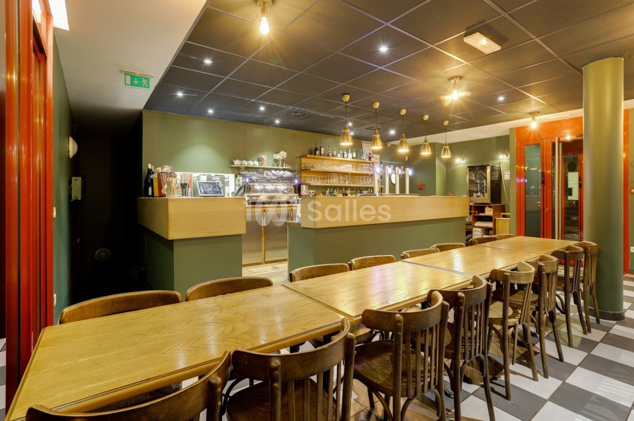 Salle de restaurant avec longues tables en bois, chaises assorties et comptoir de bar éclairé en arrière-plan.