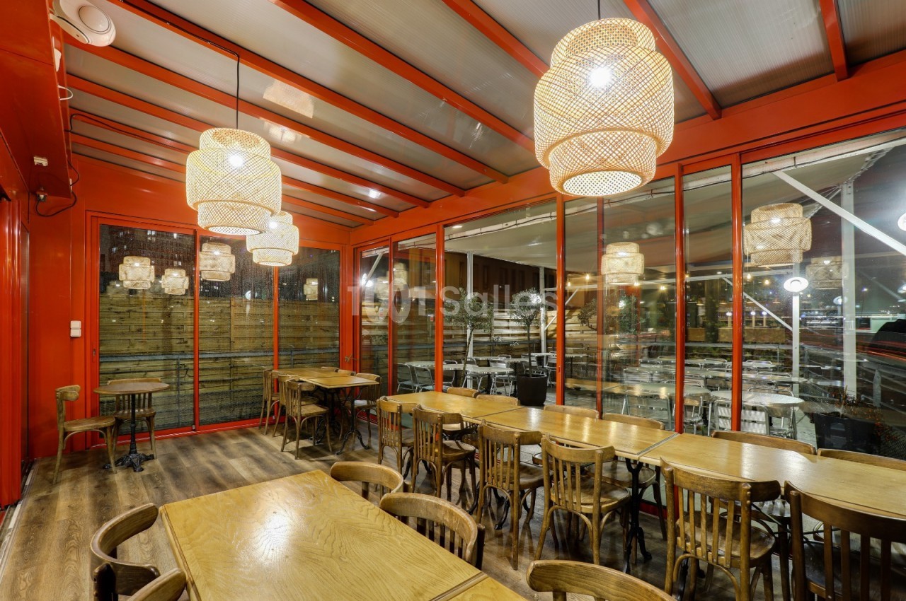Salle de restaurant avec tables en bois, chaises assorties, grandes baies vitrées et luminaires suspendus en osier.