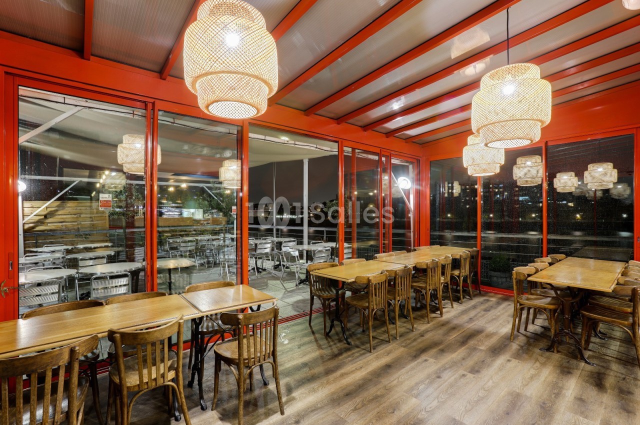 Salle de restaurant avec tables en bois, chaises assorties et grandes lampes suspendues, donnant sur une terrasse vitrée.